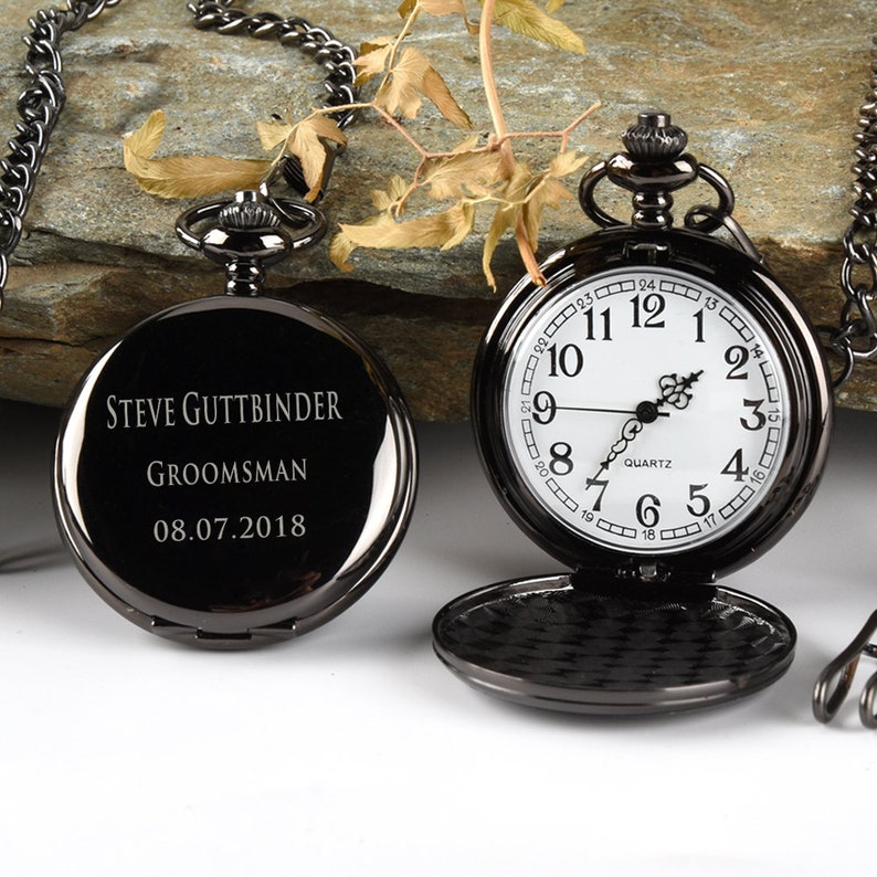 Puede incluir: Reloj de bolsillo negro con una esfera blanca y agujas negras. El reloj tiene una cadena. La parte posterior del reloj est&aacute; grabada con el texto "STEVE GUTTBINDER GROOMSMAN 08.07.2018".