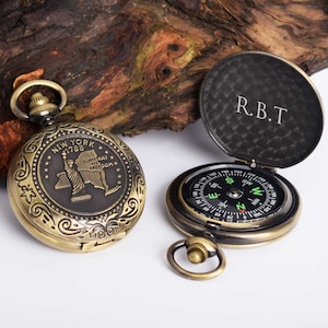 Pode incluir: Um compasso dourado com um mostrador preto e uma agulha de bússola. O compasso é gravado com as palavras "New York 1788 Gateway to Freedom" e uma representação da Estátua da Liberdade. O compasso tem uma tampa articulada com as iniciais "R.B.T." gravadas no interior.
