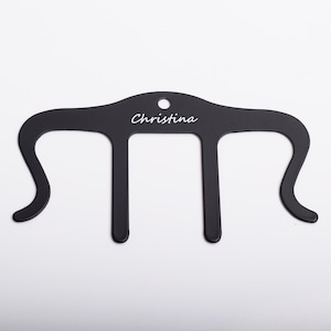 May include: Black metal hanger with three prongs and a hole at the top. The hanger is personalised with the name "Christina" in white lettering.