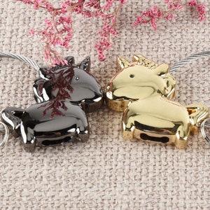 Op de afbeelding: Een paar unicorn sleutelhangers, een zwart en een gouden, met een zilveren ketting die ze verbindt. De sleutelhangers zijn scherp in beeld, en de achtergrond is een wazige beige stof.