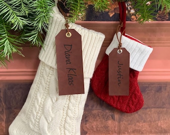 Op de afbeelding: Twee kerstsokken, één crèmekleurig en één rood, hangen aan een groene slinger. Elke sok heeft een bruin label met een naam. Op het crèmekleurige label staat "Diana Klaas" en op het rode label staat "Justin".