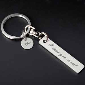 May include: A silver keychain featuring a rectangular tag inscribed with "I love you more!" and a small, round tag with the name "Rhys". The keychain includes a split ring and a lobster clasp.