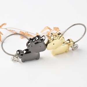 Op de afbeelding: Een zwarte en gouden metalen sleutelhanger met twee unicorn-bedels. De bedels zijn verbonden door een zilveren kabelketting. De bedels hebben de woorden "Ker" en "Amy" erop.