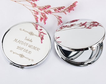 May include: A silver compact mirror with a white background and gold lettering. The mirror has the text "Isabela Maid of Honor 2017.08.10" engraved on the front.