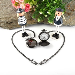 Puede incluir: Un reloj de bolsillo negro con una esfera blanca y n&uacute;meros romanos. El reloj est&aacute; unido a una cadena negra y est&aacute; sobre una superficie blanca. Dos peque&ntilde;as figuras est&aacute;n a cada lado del reloj.