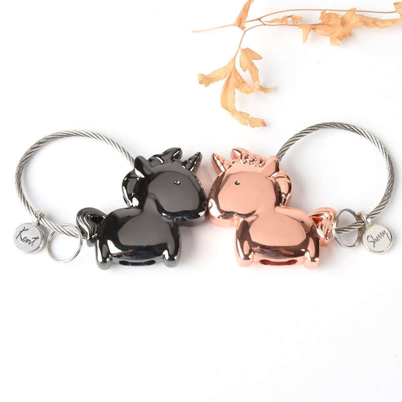 Op de afbeelding: Twee sleutelhangers in de vorm van eenhoorns, een zwart en een ros&eacute; goud, met metalen ringen en gepersonaliseerde labels met de tekst "Kent" en "Sherry".