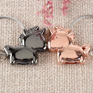 Op de afbeelding: Twee sleutelhangers in de vorm van eenhoorns, een zwart en een ros&eacute; goud, zijn bevestigd aan zilveren metalen kettingen. De eenhoorns kijken elkaar aan en lijken elkaar te kussen.