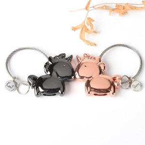 Op de afbeelding: Twee sleutelhangers in de vorm van eenhoorns, een zwart en een ros&eacute; goud, met metalen ringen en gepersonaliseerde labels met de tekst "Kent" en "Sherry".