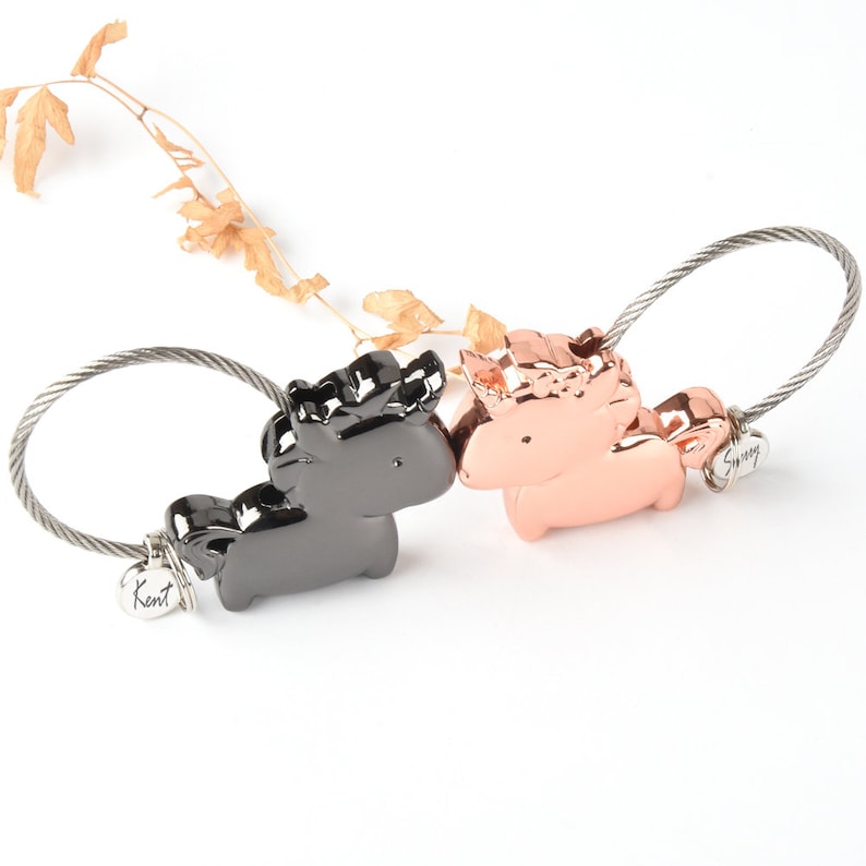 Op de afbeelding: Een zwarte en ros&eacute;gouden metalen sleutelhanger met twee unicorn-bedels. De sleutelhanger heeft een zilveren kabellus en een klein label met de tekst "Kent".