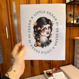 May include: A rectangular art print featuring a central illustration of a person with dark wavy hair, surrounded by yellow stars and teal flowers. The text "DREAM A LITTLE DREAM OF ME" is printed in a circular pattern around the illustration.