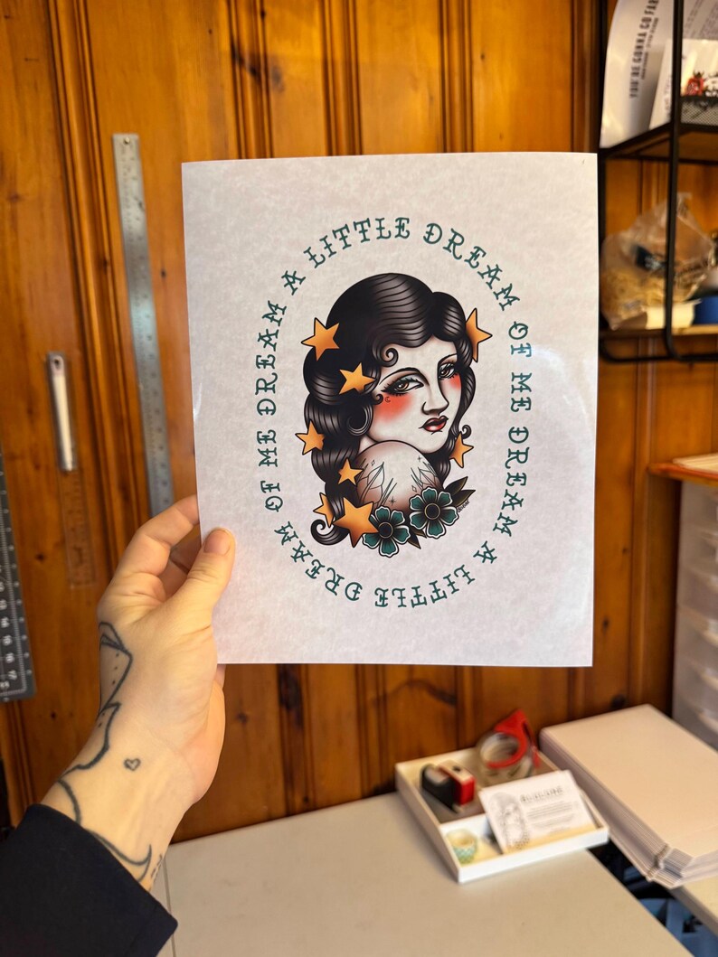 May include: A rectangular art print featuring a central illustration of a person with dark wavy hair, surrounded by yellow stars and teal flowers. The text "DREAM A LITTLE DREAM OF ME" is printed in a circular pattern around the illustration.