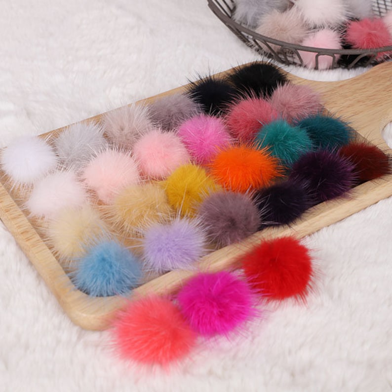 May include: A wooden tray filled with a variety of colorful, fluffy pom-poms. The pom-poms are in shades of pink, orange, yellow, blue, purple, and black. They are arranged on a light-colored, textured surface.