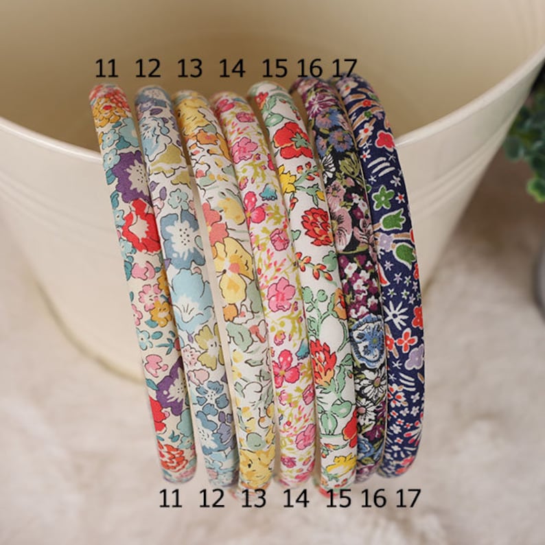 K&ouml;nnte beinhalten: Sieben Haarreifen mit floralen Mustern. Jeder Haarreifen ist mit einem Stoff mit verschiedenen Blumenmustern bedeckt, darunter rote, rosa, gelbe, blaue und violette Blumen. Die Haarreifen sind mit den Nummern 11 bis 17 versehen.