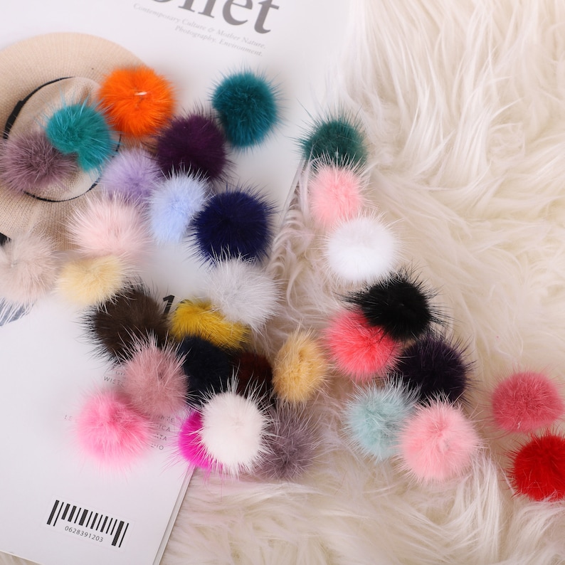 May include: A collection of colorful, fluffy pom-poms in various shades, including pink, orange, green, and purple. The pom-poms are scattered on a white, textured surface, suggesting a craft or accessory theme. Some are resting on a beige beret.