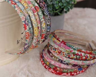 May include: A collection of floral patterned headbands in various colors, including red, pink, blue, and yellow. The headbands are stacked and arranged on a white surface, with a few leaning against a white bucket. The headbands have a fabric covering.