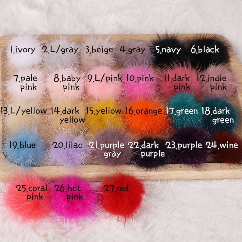 May include: A collection of 27 fluffy pom-poms in various colors, including ivory, gray, beige, navy, black, pink, yellow, orange, green, blue, purple, and red. Each pom-pom is labeled with a number and color name.