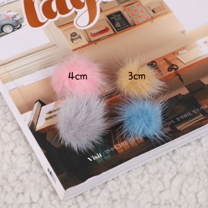 May include: Four fluffy pom-poms in pink, yellow, gray, and blue, with sizes labeled as 4cm and 3cm. The pom-poms are on a magazine page with text that reads "Visit" and other Korean text.