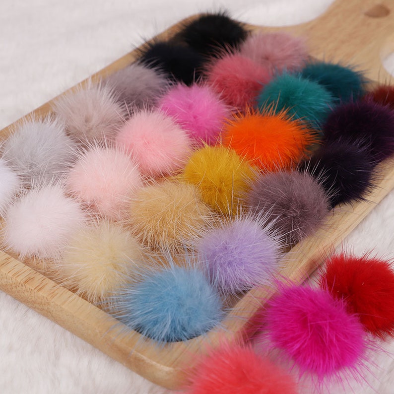May include: A wooden tray filled with a variety of colorful, fluffy pom-poms. The pom-poms are in shades of pink, blue, yellow, orange, purple, and green. The texture of the pom-poms appears soft and furry, suggesting they are made of fur.