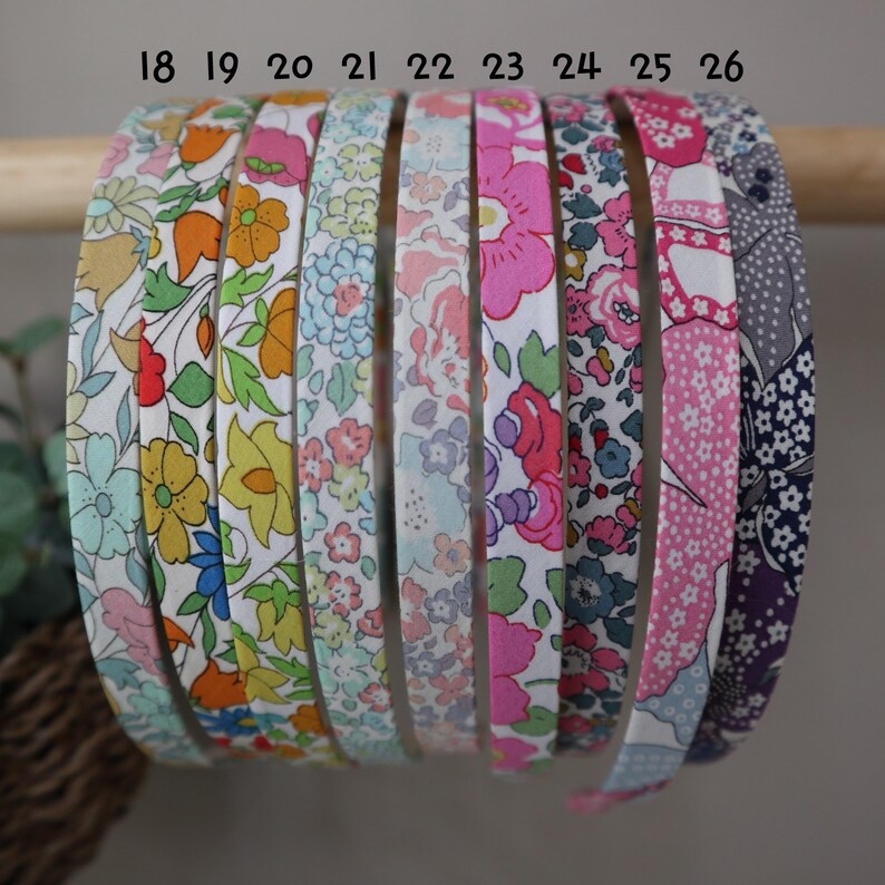 K&ouml;nnte beinhalten: Eine Auswahl an Haarreifen mit floralen Mustern in verschiedenen Farben, darunter Rosa, Blau, Gelb und Orange. Die Haarb&auml;nder werden auf einem Holzstab pr&auml;sentiert, der die verschiedenen Stoffdesigns hervorhebt. Die Nummern 18 bis 26 sind auf den Haarb&auml;ndern aufgedruckt.