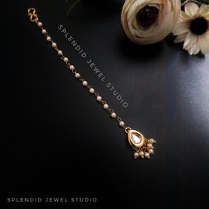 May include: A gold nose ring with a teardrop-shaped stone and a chain of white pearls. The chain is attached to a hook at the top. The text "SPLENDID JEWEL STUDIO" is visible on the image.