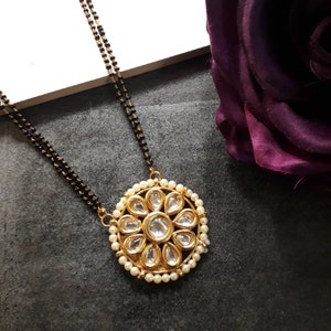 May include: A gold-toned pendant necklace with a floral design. The pendant features clear, teardrop-shaped stones surrounded by small pearls. The necklace has a black beaded chain.