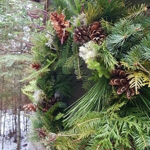 K&ouml;nnte beinhalten: Ein gr&uuml;ner Weihnachtskranz mit Kiefernnadeln, Zeder und Tannenzapfen. Der Kranz ist mit getrockneten Blumenzweigen und wei&szlig;em Flechten verziert.