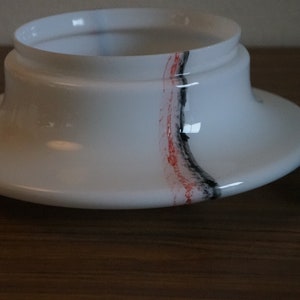 K&ouml;nnte beinhalten: Wei&szlig;er Lampenschirm aus Glas mit einem schwarzen und roten Streifenmuster. Der Lampenschirm ist von seinem Sockel getrennt.