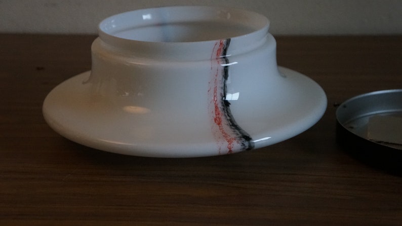 K&ouml;nnte beinhalten: Wei&szlig;er Lampenschirm aus Glas mit einem schwarzen und roten Streifenmuster. Der Lampenschirm ist von seinem Sockel getrennt.
