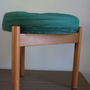 May include: A green foam-covered stool with a wooden frame and four legs. The stool is sitting on a brown wooden surface.