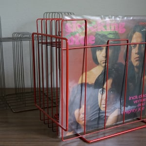 May include: Two wire record holders. One is red and holds a vinyl record with a band photo. The other is silver. Both are made of thin metal rods and sit on a wooden surface. The red holder is in focus.