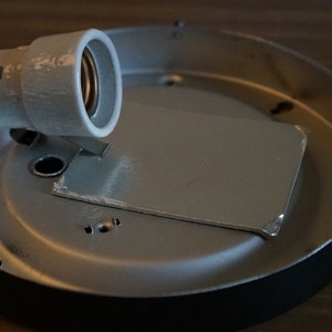 K&ouml;nnte beinhalten: Eine silberne Deckenleuchte aus Metall mit einer wei&szlig;en Kunststoff-Lampenfassung und einer Metall-Montageplatte.