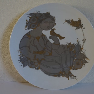 May include: A white ceramic plate with a gold and gray design of a woman with birds. The woman is sitting in a floral setting.