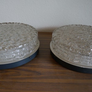 Puede incluir: Dos lámparas de techo vintage de vidrio transparente con un patrón texturizado en forma de burbujas. Las lámparas tienen una base de metal gris oscuro y se exhiben sobre una superficie de madera. Las lámparas son redondas y de diseño retro.