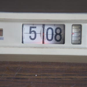 May include: Cream-colored vintage flip clock displaying "5:08". The clock has a rectangular shape with a clear plastic front. A small dial on the right side shows the minutes. The brand name "Copal" is visible on the left side.