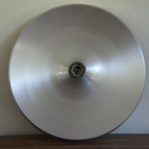 May include: A circular, brushed silver metal light fixture shade with a smooth, concave surface. The center features a dark, circular socket opening designed to hold a light bulb, creating a clean, reflective, and metallic appearance.