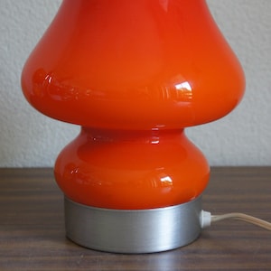 Op de afbeelding: Een retro oranje paddenstoellamp met een afgeronde boven- en onderkant. De lamp heeft een glanzende afwerking en staat op een zilverkleurige voet. Een wit elektrisch snoer komt uit de voet.