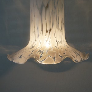 Peut inclure: Un luminaire suspendu en verre blanc avec un bord festonné et un motif marbré. La lumière est allumée et diffuse une lueur chaude.