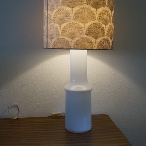 May include: A white ceramic table lamp with a brown and white patterned lampshade. The lamp is turned on and illuminating the room.