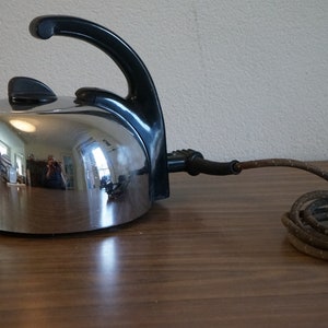 Può includere: Un bollitore elettrico cromato con manico nero e cavo marrone. Il bollitore è su una superficie di legno.