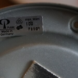 K&ouml;nnte beinhalten: Ein silberner Lampenfu&szlig; aus Metall mit einem Etikett mit der Aufschrift "max. Watt 100", "DE", "Putzler", "Made in Germany" und "761101".