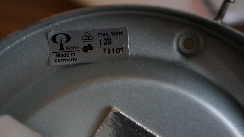 K&ouml;nnte beinhalten: Ein silberner Lampenfu&szlig; aus Metall mit einem Etikett mit der Aufschrift "max. Watt 100", "DE", "Putzler", "Made in Germany" und "761101".