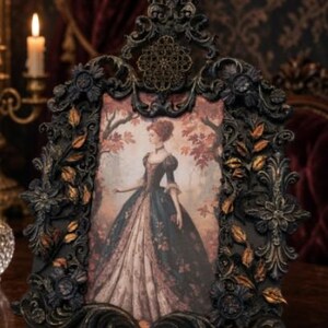 May include: A rectangular picture frame with an ornate, dark, distressed finish featuring sculpted floral and leaf motifs with gold highlights. The frame holds an illustration of a person in a long, dark gown standing in a forest.