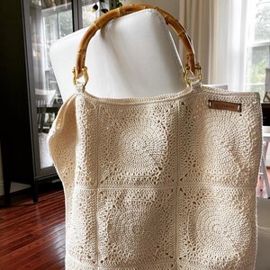 May include: A crocheted tote bag with a square pattern and a bamboo handle. The bag is cream colored and has a small leather tag.