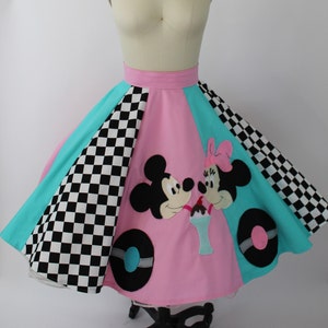 May include: A colorful skirt featuring Mickey and Minnie Mouse. The skirt has a pink center panel with the characters, flanked by turquoise and black and white checkered panels. Black and white tire-shaped accents are on the pink panel.