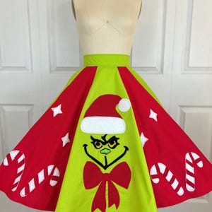 May include: A festive Christmas skirt featuring a Grinch design. The skirt has a lime green center panel with the Grinch's face and a red bow. The outer panels are red with white candy canes and stars, and a lime green waistband.