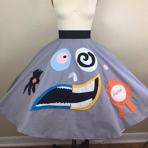 May include: A gray skirt featuring a black waistband and various felt appliques including a light blue eye, a white spiral eye, a blue and white nose, a wide mouth with teeth, a black shape, and an orange badge with the word MAYOR written on it.