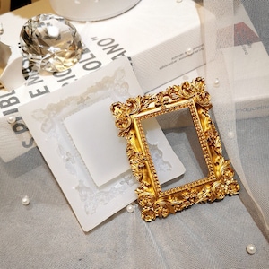 May include: A gold-colored ornate picture frame with a white silicone mold in the background. The mold is shaped like a picture frame.