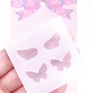 May include: A clear silicone mold featuring two wing and two butterfly shapes. The mold is held in a hand, with a pink bow-shaped tag in the background. The tag has a diamond pattern and white polka dots.
