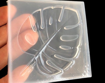 May include: A clear, square silicone mold featuring a detailed monstera leaf design. The mold is held against a dark background, showcasing the leaf's intricate veins and shape. Ideal for crafting resin art or other DIY projects.