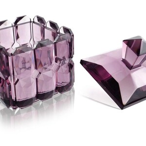 May include: A decorative, faceted, amethyst-colored glass box with a matching lid. The box is rectangular with a geometric design, and the lid features a square top with a diamond-shaped handle.
